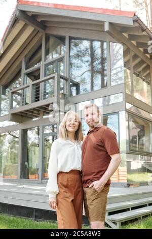 Portrait of smiling young couple in casual outfits standing against glass house and embracing each other Stock Photo