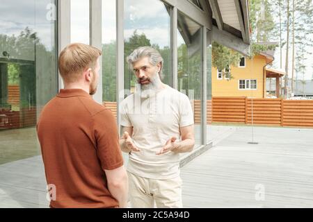 Senior gray-haired father standing porch of cottage house and gesturing hands while talking to adult son Stock Photo
