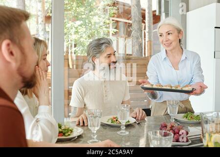 Smiling careful senior mother giving appetizers to family while they having dinner together in dining room Stock Photo