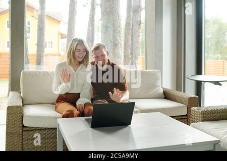 Cheerful young couple sitting on sofa and waving hands to laptop while chatting via video link in living room of cottage house Stock Photo