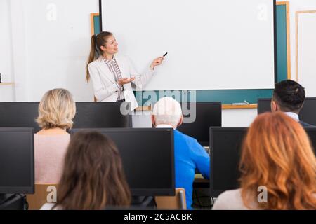 Friendly young female teacher lecturing attentive adult students of different ages in computer room Stock Photo