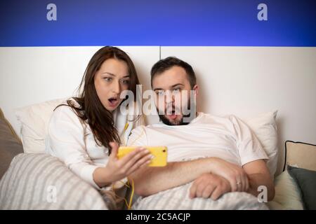 Shocked and scared couple watching horror movies on smartphone lying in bed Stock Photo