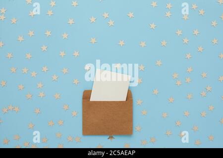 white blank card in kraft brown paper envelope on blue background with confetti. Holiday mockup. Top view. Stock Photo