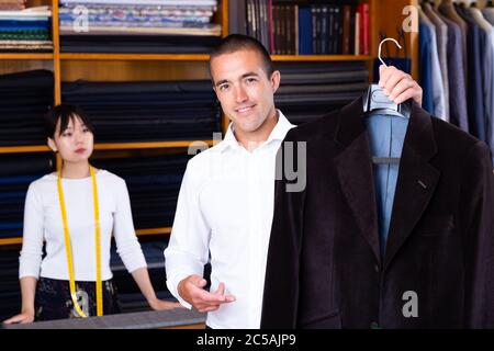 Satisfied male client of sewing workshop showing his new suit jacket on background of young Chinese female tailor Stock Photo