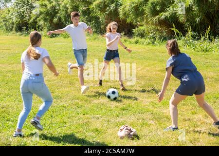 Happy teenage girls and guy playing football on green lawn in summer city park Stock Photo
