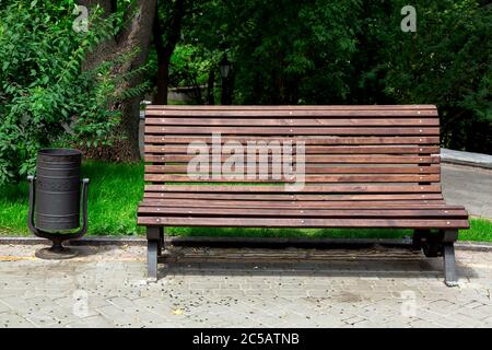 a wooden bench made of brown boards on the back and an iron black litter bin on a stone sidewalk in a park with trees and green grass, empty seat nobo Stock Photo