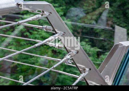 a corner of a glass bridge with tension steel cables and a cable tensioner with thread over green trees. Stock Photo