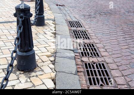 hatches of the drainage system of the road paved with paving stones with a curb and stone tiles on the sidewalk with iron columns on chains for the sa Stock Photo