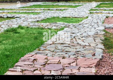 footpaths made of natural rough stone paved with mosaic in the lawn grass in the old town. Stock Photo