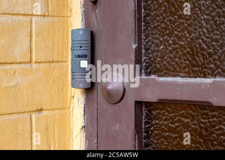intercom with a doorbell and video link, closeup of a security device on a yellow brick wall near a wooden door with glass on outdoor nobody. Stock Photo