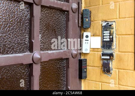 several types of different intercoms and electronic locks on a chip or card on a yellow brick wall on outdoor against a wooden door with glass inserts Stock Photo
