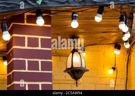 iron hanging on the wooden ceiling retro lantern with a light bulb on the brick facade of a building decorated with a garland with light bulbs, close- Stock Photo