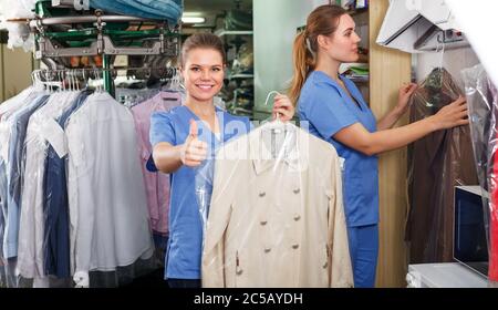 Two confident smiling female workers offering professional dry cleaning, showing clean clothing Stock Photo
