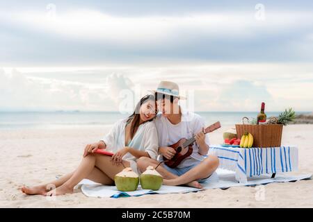 Asian couple happy and enjoy the trip honeymoon of couple lover on the sea beach by playing Ukulele and singing song music together. Summer, holidays, Stock Photo