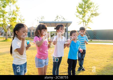 Large group of happy Asian smiling kindergarten kids friends playing blowing bubbles together in the park on the green grass on sunny summer day. Stock Photo