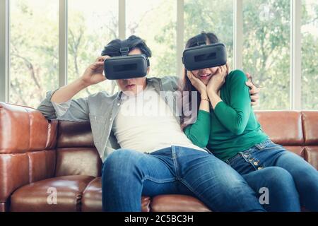 Young Couple Having Fun While Watching Video Via Virtual Reality Together. Couple Love Having Enjoyment With Electronic VR Goggles Gaming on Couch. En Stock Photo