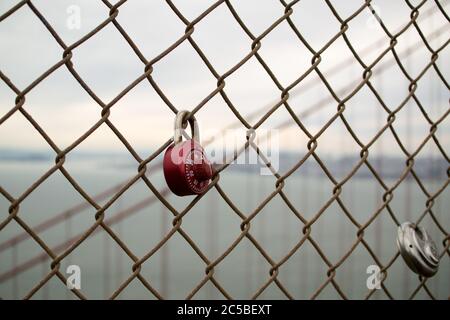 Love locks on Golden Gate Bridge, one red combination lock, one round padlock. San Francisco skyline in the background. Grey sky. Stock Photo