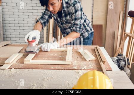 Carpenter Man is Working Timber Woodworking in Carpentry Workshops, Craftsman is Using Sander Machine on Timber Frame for Wooden Furniture in Workshop Stock Photo