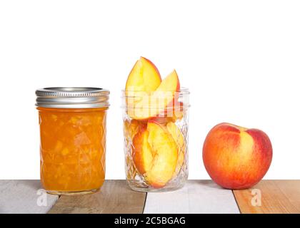 Fresh sliced peaches in an open mason jar with whole peach on table next to a full jar of home made canned peach jam on light colored wood table, isol Stock Photo