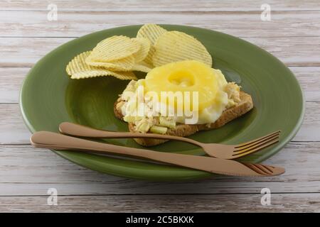 Open face tuna salad sandwich with melted provolone cheese slice and pineapple fruit ring with potato chips on green plate with knife and fork Stock Photo