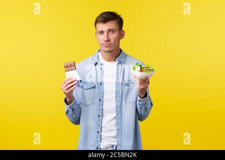 Healthy lifestyle and people emotions concept. Gloomy handsome caucasian guy showing salad in bowl and candy bar, grimacing as reluctant eat diet food Stock Photo