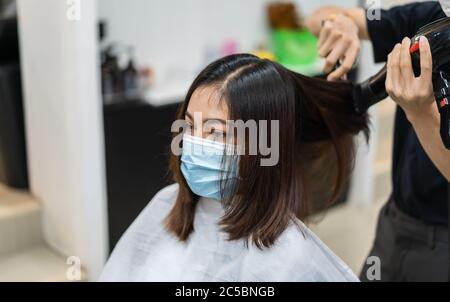 hairdresser using hair dryer and comb to the hair of woman, people must be wering a medical mask to protection coronavirus (covid-19) pandemic. new no Stock Photo