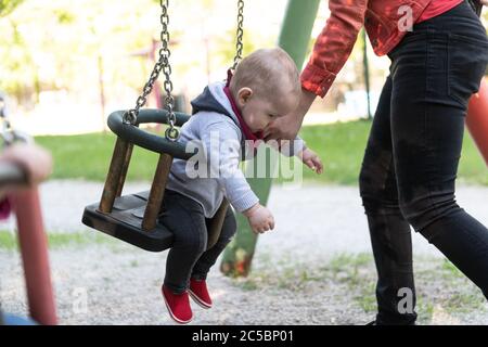 Happy Vivacious Young Mother With Her Baby Son Playing Together Outdoors in a Playground Pushing Him on a Swing as He Laughs Happily Stock Photo
