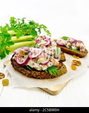 Salmon, petiole celery, raisins, walnuts, red onions and curd cheese salad on toasted bread with green lettuce leaves on parchment on a wooden board b Stock Photo