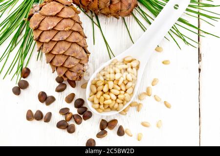 Peeled kernels of cedar nuts in a spoon, two pinecones and nuts in the shell against a light wooden board on top Stock Photo