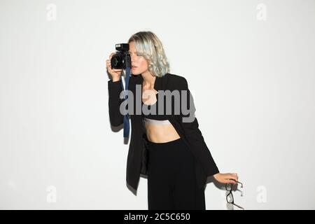 Young lady in black classic costume thoughtfully taking photos on retro camera with eyeglasses in hand over white background isolated Stock Photo