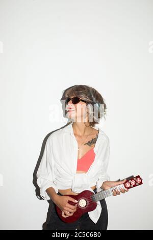 Cool smiling girl in white shirt and sunglasses happily looking aside while playing on little guitar over white background Stock Photo