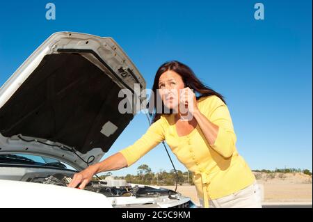 Stressed mature woman breakdown with car on remote road calling for assistance, for help on mobile phone, isolated with blue sky as background and cop Stock Photo