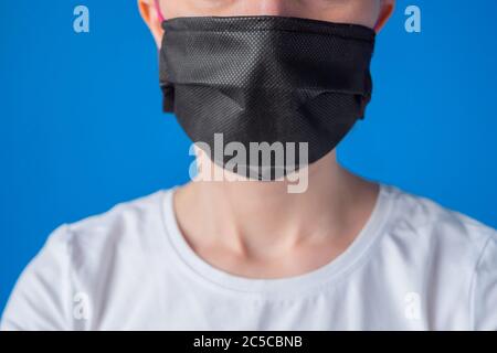 Portrait of unrecognizable woman wearing black medical face mask, white shirt in room with blue wall at home. Self isolation, prevention, quarantine Stock Photo