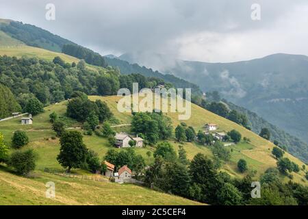 Scattered farm houses on a green hill  dotted with Alpine pastures, at a cloudy rainy day, region Lombardy, Italy. Italian rural summer landscape. Stock Photo