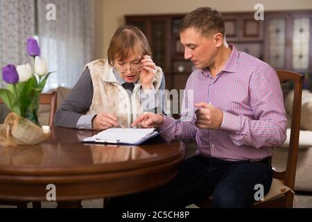 Adult man with his elderly mother discussing documents while sitting at home table Stock Photo
