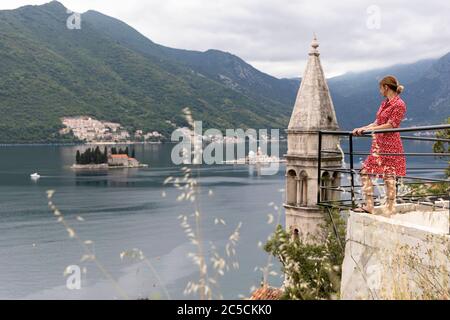 woman in red dress standing at fence at viewpoin with scenic panorama view of the historic town of Perast at famous Bay of Kotor, Montenegro Stock Photo