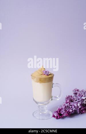 Beautiful dalgona drink a frothy coffee in a transparent mug and flowers lilac to purple Il purple background. Next to the mug is a marshmallow and a Stock Photo