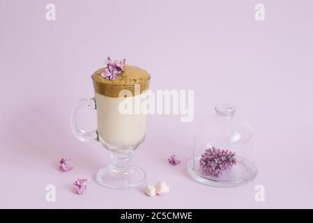 trendy drink 2020 dalgon Coffee in a transparent Cup with a paper tube, a plate with lilac flowers on a lilac background with a copy space Stock Photo