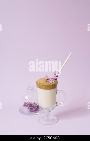 dalgona Coffee in a transparent Cup with a paper tube, a plate with lilac flowers on a lilac background with a copy space Stock Photo