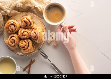 cropped view of woman holding cup of coffee near homemade cinnamon rolls on marble surface with fork, condensed milk and cloth Stock Photo
