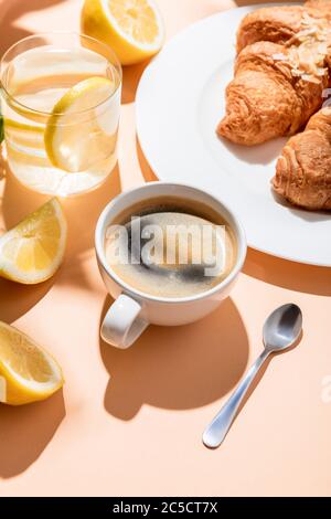 coffee cup with teaspoon, croissants and glass of water with lemon for breakfast on beige table Stock Photo