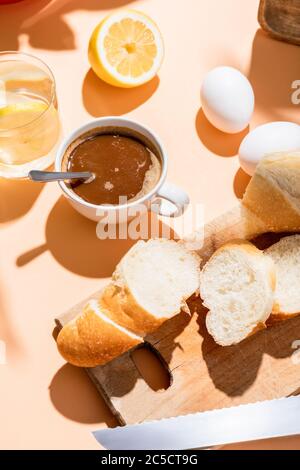 coffee cup with teaspoon, eggs, baguette and glass of water with lemon for breakfast on beige table Stock Photo