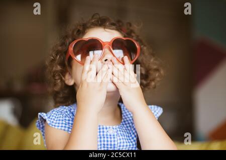 Portrait of pretty baby girl with dark curly hair in sunglasses dreamily covering mouth with hands at home Stock Photo