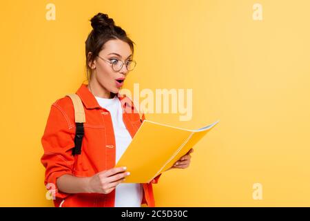 shocked student in glasses looking at copy book isolated on yellow Stock Photo
