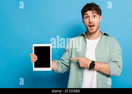 shocked young man pointing at digital tablet with blank screen on blue Stock Photo