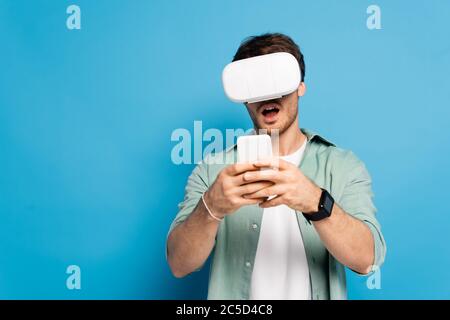 shocked young man in vr headset using smartphone on blue Stock Photo