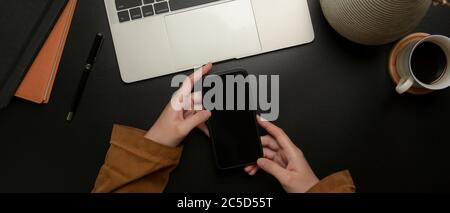Overhead shot of hands holding smartphone on dark modern workspace with laptop, supplies and decoration Stock Photo