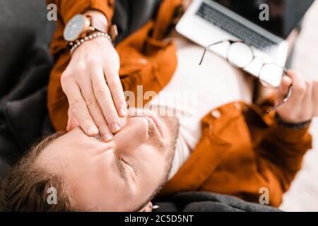 Overhead view of tired 3d designer touching eye and holding eyeglasses with laptop on couch Stock Photo