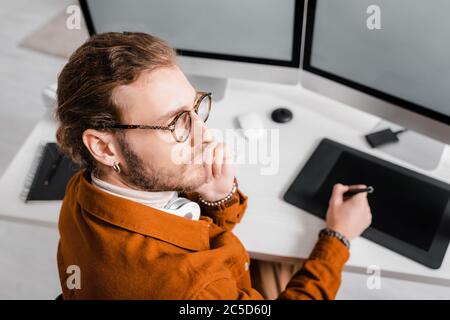 Selective focus of pensive 3d artist looking away while working with graphics tablet and computers at table Stock Photo