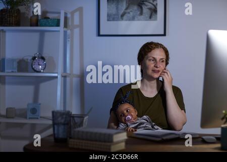 Business mother using computer and talking on mobiloe phone while sitting at the table with baby on her knees Stock Photo
